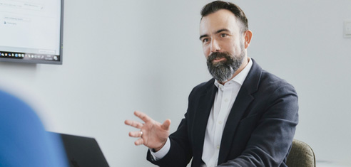 Ein bärtiger Mann in einer Anzugsjacke sitzt an einem Schreibtisch und spricht mit der Hand, vor ihm steht ein Laptop, im Hintergrund ist ein Bildschirm zu sehen.