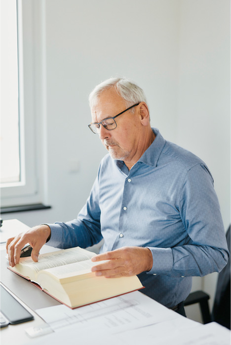 Ein älterer Mann mit grauem Haar und Brille sitzt an einem Schreibtisch und liest ein großes aufgeschlagenes Buch. Er trägt ein hellblaues Hemd und scheint konzentriert zu sein, während er in der Nähe Papiere und ein Fenster sieht.