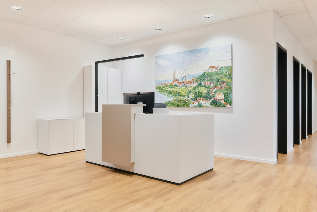 Ein moderner, minimalistischer Empfangstresen in einem hellen Büro mit hellem Holzfußboden, weißen Wänden und einem großen bunten Landschaftsgemälde hinter dem Tresen. Mehrere Türen säumen den Korridor auf der rechten Seite.