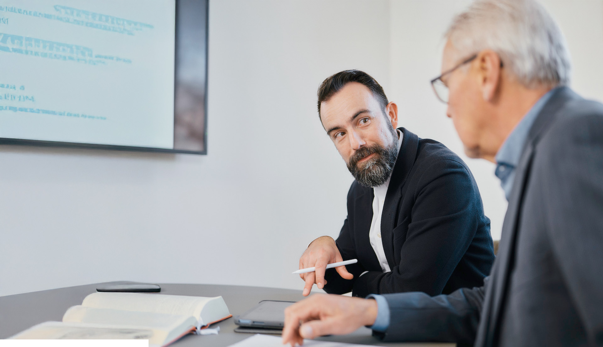 Zwei Männer in Geschäftskleidung sitzen an einem Tisch mit aufgeschlagenen Büchern und Dokumenten und führen ein Gespräch. Ein bärtiger Mann schaut direkt in die Kamera, während der andere ältere Mann ihm zugewandt ist. Im Hintergrund ist ein Bildschirm mit Text zu sehen.