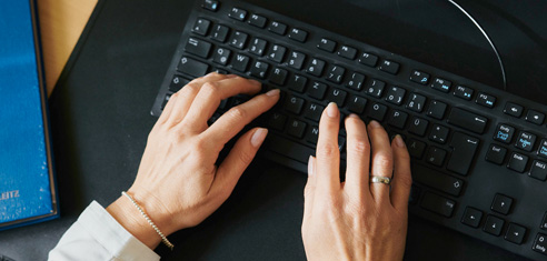 Eine Person mit einem Ring und einem Armband tippt auf einer schwarzen Computertastatur, in deren Nähe ein blauer Ordner und eine Computermaus zu sehen sind.