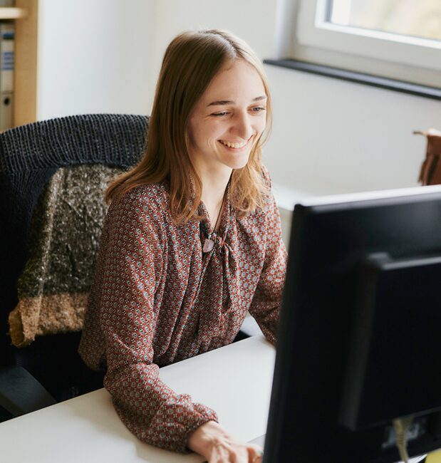 Eine Frau mit langen braunen Haaren und einer gemusterten Bluse sitzt an einem Schreibtisch und arbeitet lächelnd an einem Computer in einem hellen Büro.