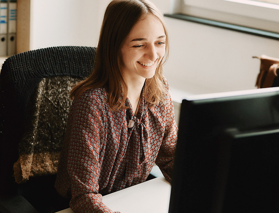 Eine Frau mit braunen Haaren und einer gemusterten Bluse sitzt an einem Schreibtisch und schaut lächelnd auf einen Computermonitor in einer hellen Büroumgebung.